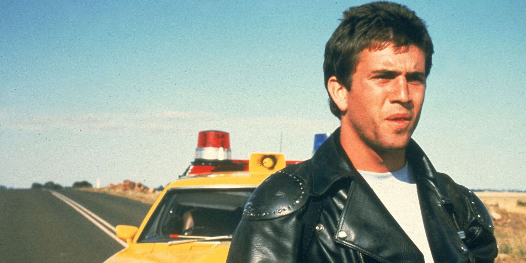 Mel Gibson con una chaqueta de cuero negra y una camiseta blanca parado detrás de su coche de policía amarillo.