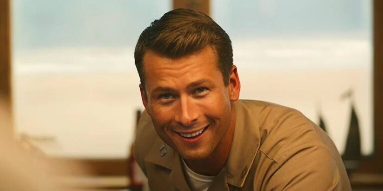 Close -up of actor Glen Powell smiling, wearing a brown naval uniform like Jake in Top Gun: Maverick